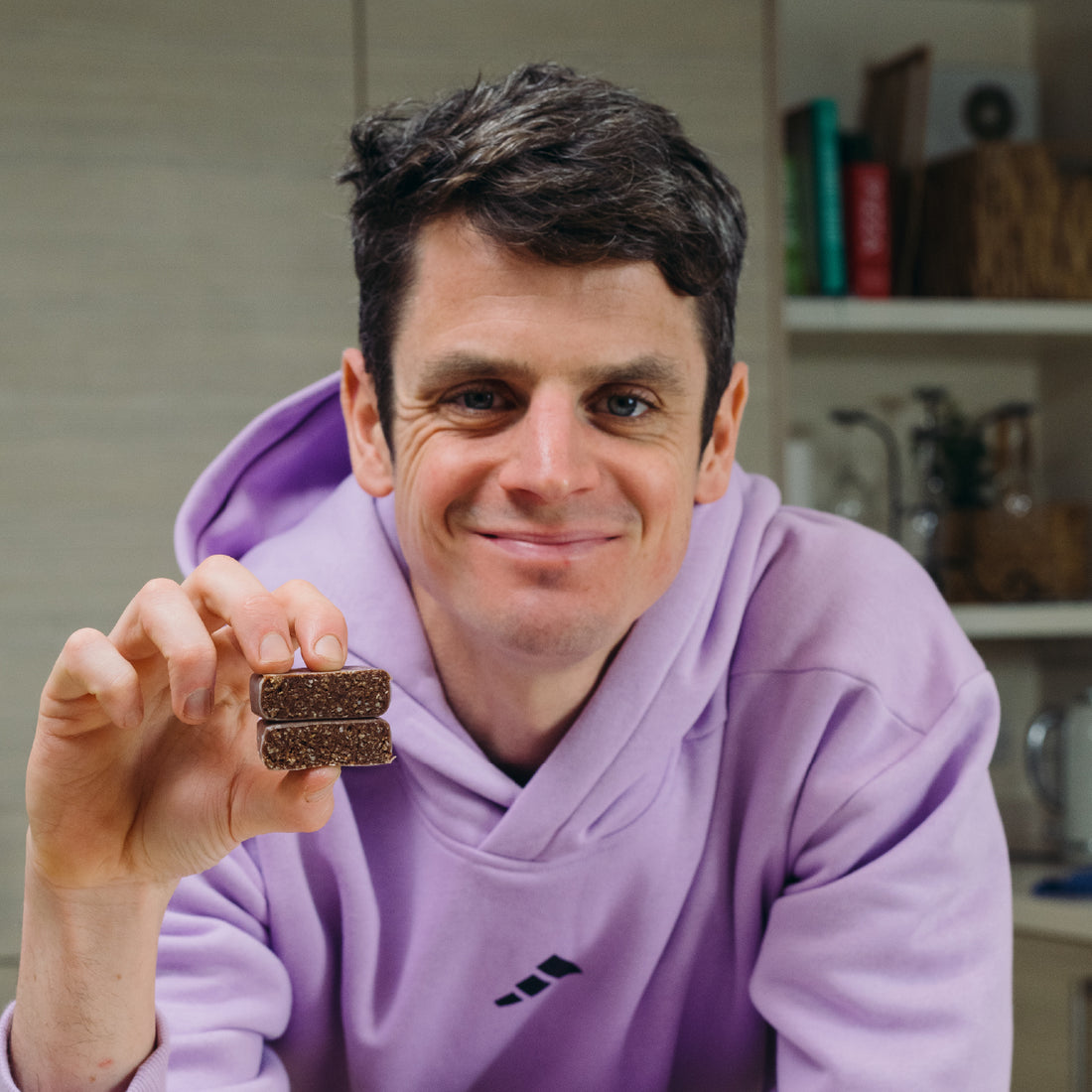Jonny Brownlee in a purple hoodie holding a Chocolate Brownlee OGT bar in a kitchen setting
